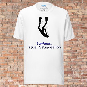 以下が含まれることがあります： 白いTシャツ。黒いシルエットのダイバーとフィンが描かれています。「Surface... Is Just A Suggestion」というテキストが青色でプリントされています。Tシャツはクルーネックで半袖です。
