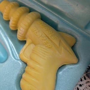 May include: A yellow, gun-shaped soap bar with the embossed text BUBBLE BLAZER and AVON on the surface. The soap is resting inside a light blue, molded plastic container that matches the shape of the toy-like design of the soap bar.
