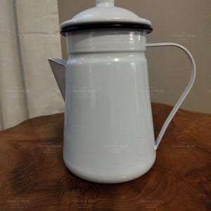 May include: A white enamel coffee pot with a matching lid, a curved handle, and a spout. The pot features a smooth, glossy finish with a thin black rim around the lid and the top edge of the vessel, presenting a clean and functional design.