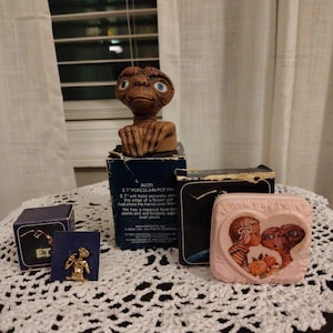May include: A collection of E.T. the Extra-Terrestrial memorabilia including a brown porcelain figurine, a small gold-toned pin on a blue card, and a square pink trinket box featuring a heart-shaped illustration of a child and E.T.