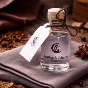May include: A clear glass reed diffuser bottle with a wooden stopper and a tag. The label reads "Leather & Tobacco" and "Moon Mist". The bottle is tied with twine and sits on a grey cloth.