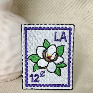 May include: Embroidered patch featuring a white magnolia flower with green leaves, a brown center, and the text "LA" in purple. The patch has a purple border and the text "12¢".