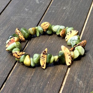 May include: A beaded bracelet featuring a mix of green and tan-colored stones. The bracelet is made of various shades of green stones, with some stones having a tan and brown pattern. The bracelet is displayed on a dark wooden surface.