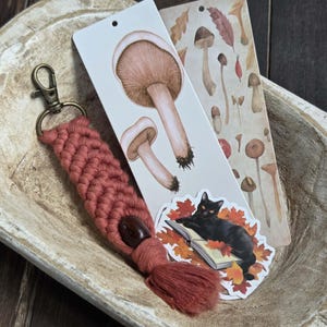 Puede incluir: Un llavero de macramé en color rojo-naranja apagado con una cuenta de madera y borla. Dos marcadores presentan ilustraciones de hongos y un gato negro leyendo un libro rodeado de hojas de otoño. Los artículos están dispuestos en un cuenco de madera.
