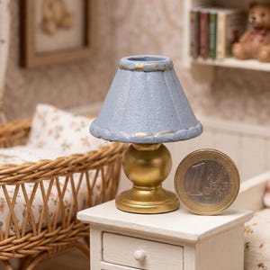 Op de afbeelding: Miniatuur blauwe lamp met een gouden voet en rand, op een wit nachtkastje. Een één euro munt ligt naast de lamp. De scène suggereert een poppenhuis slaapkamer, met een rieten wieg en ingelijste kunstwerken op de achtergrond.
