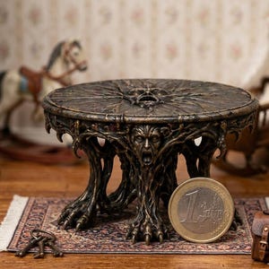 May include: A miniature bronze-colored table with a detailed, textured design. The round tabletop features a face in the center, and the legs are sculpted to resemble tree roots. A Euro coin is next to the table for scale. A set of keys and a small rug are also visible.