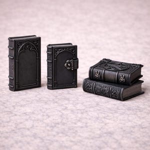 Puede incluir: Una colección de libros en miniatura de estilo gótico, de color gris oscuro. Los libros presentan intrincados diseños en relieve, incluyendo marcos arqueados, cierres florales y un pentagrama. Algunos están apilados.