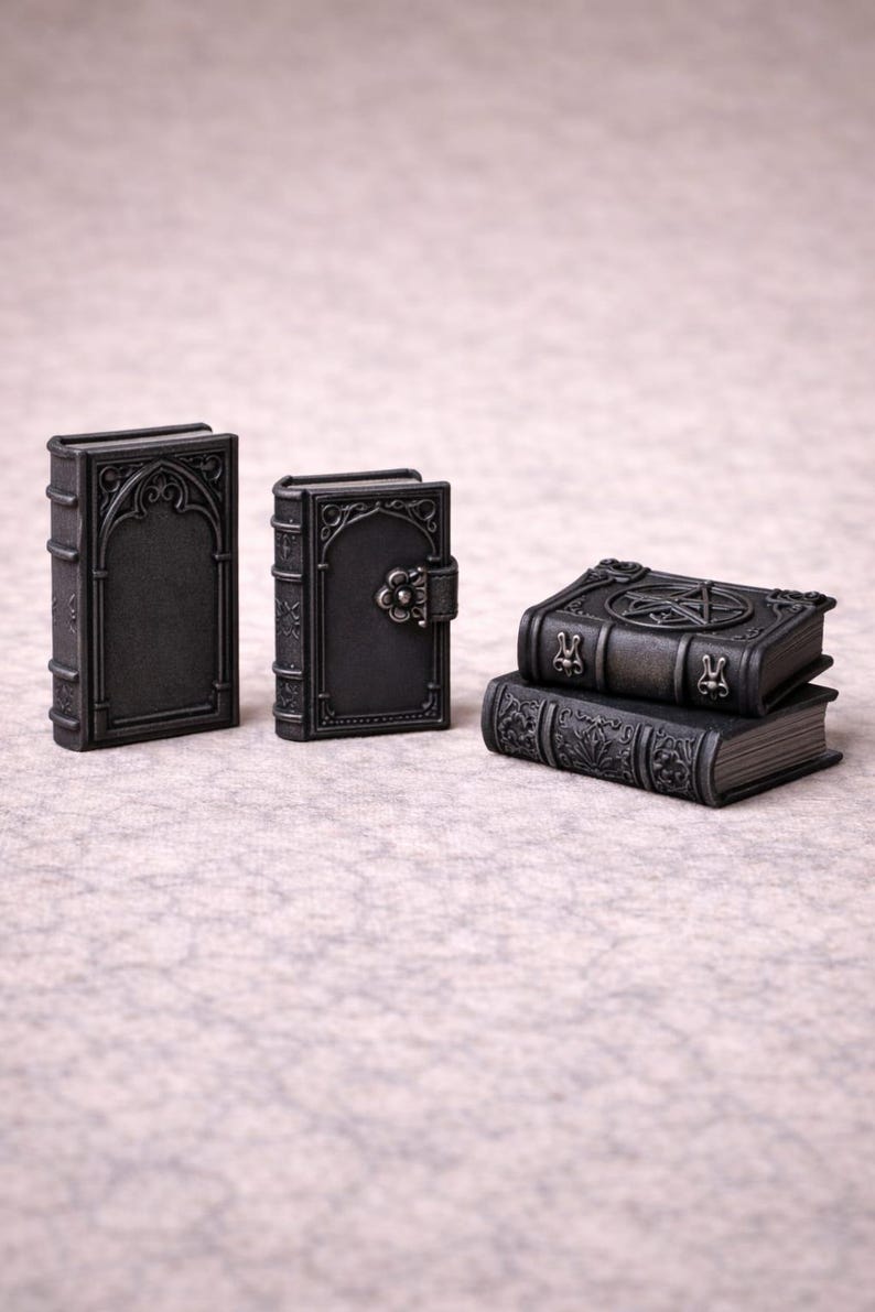 May include: A collection of miniature, dark gray, gothic-style books. The books feature intricate embossed designs, including arched frames, floral clasps, and a pentagram. Some are stacked, showcasing detailed spine patterns.
