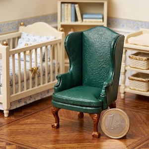 May include: Miniature green leather wingback chair with ornate legs, set in a dollhouse nursery. The chair is next to a crib and a changing table. A Euro coin is next to the chair.