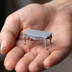 May include: A miniature gray table with ornate detailing, held in a person's hand. The table has a rectangular top and curved legs, with decorative carvings along the apron. This dollhouse furniture piece is a charming addition to any miniature collection.