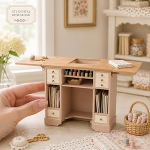 May include: A miniature wooden sewing desk with a light wood top and a soft pink base. The desk features multiple small drawers with gold knobs, open shelving for thread spools, and vertical storage for books or fabric. Text reads STL DIGITAL DOWNLOAD.