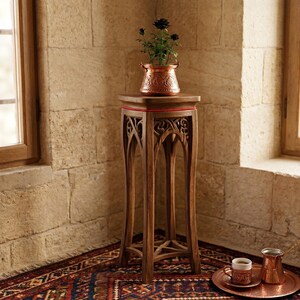 Peut inclure: Un support de plante en bois sculpté avec un pot en cuivre contenant une plante sombre. Le support est placé dans une pièce aux murs en pierre et sur un tapis à motifs.