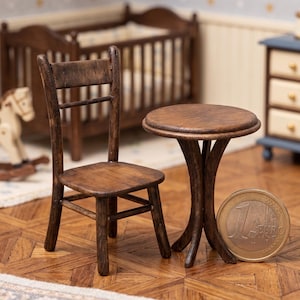 Peut inclure: Ensemble miniature de chaise et de table en bois. La chaise a un dossier à lattes et une assise carrée. La table a un plateau rond et un piètement incurvé à trois pieds. Une pièce d'un euro est à côté de la table. L'ensemble est en bois brun foncé.