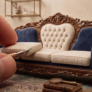 May include: Miniature ornate sofa with a cream-colored button-tufted back and seat cushions. The sofa has dark blue side cushions and armrests, and a dark brown, intricately carved wooden frame. A hand holds a tool near a cushion.