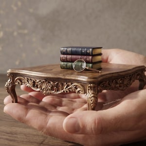 May include: A miniature ornate wooden table with three small books and a magnifying glass. The table has detailed carvings and is held in open hands. The books are bound in blue, burgundy, and green.