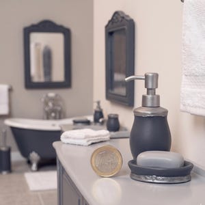 May include: A dark gray soap dispenser with a silver pump and trim, next to a matching soap dish holding a bar of soap. A one euro coin is on the counter. The bathroom has a dark gray mirror and bathtub.
