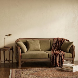 Może przedstawiać: Sofa w stylu vintage w kolorze oliwkowym z trzema pasującymi poduszkami i wzorzystym bordowym pledem. Drewniana rama ma ozdobne rzeźbienia. Po lewej stronie znajduje się mały stolik boczny z mosiężną lampą i książkami.