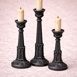 May include: Three black gothic-style candle holders with cream-colored candles. The candle holders have ornate detailing and varying heights. The candles have a melted wax appearance, adding to the vintage aesthetic.