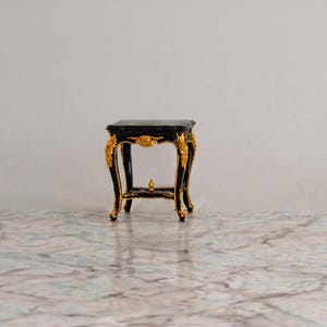 Könnte beinhalten: Ein kleiner, verzierter schwarzer Tisch mit goldenen Akzenten. Der Tisch hat eine quadratische Oberseite und geschwungene Beine mit aufwendigen goldenen Details. Der Tisch ist ein dekoratives Stück, möglicherweise zur Präsentation oder als Beistelltisch. Ca. 30 cm x 30 cm.