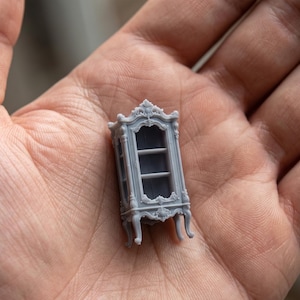 May include: A miniature gray cabinet with ornate detailing, including curved legs and a decorative top. The cabinet has a glass door and two shelves inside. The piece is held in a person's hand, showcasing its small size.