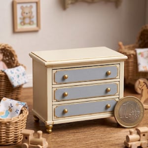 May include: Miniature cream-colored wooden dresser with three gray drawers, gold accents, and gold knobs. The dresser is part of a dollhouse set, with a framed bear picture and a toy train nearby. A Euro coin is next to the dresser.