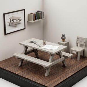 Op de afbeelding: Een miniatuur grijze picknicktafel met aangehechte banken, een stoel en een ingelijste foto. Een schuifmaat, een spoel garen en een tekening liggen op de tafel. De scène is geplaatst op een houten oppervlak.
