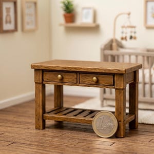 Puede incluir: Una mesa de madera en miniatura con dos cajones y un estante inferior. La mesa es de color marrón cálido con pomos dorados. Se coloca una moneda de un euro junto a la mesa para la escala.