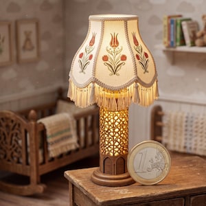May include: A miniature lamp with a cream-colored lampshade adorned with red floral designs and fringe. The lamp has a decorative, openwork base and a wooden stand. A one Euro coin is next to the lamp, suggesting the lamp's small size.