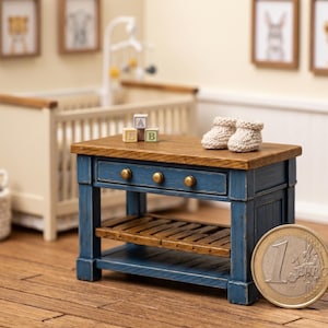 Peut inclure: Une petite table en bois bleu et marron avec un tiroir et une étagère, surmontée de blocs de bois et de chaussons de bébé tricotés. Une pièce d'un euro est à côté de la table. La table est dans une chambre d'enfant avec un berceau et des œuvres encadrées.