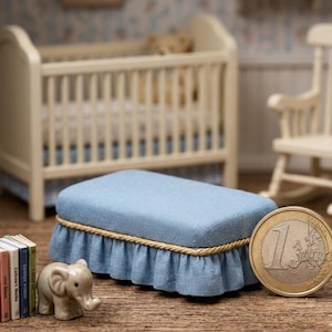 May include: Miniature blue upholstered ottoman with a ruffled skirt and gold rope trim. A small ceramic elephant and a stack of miniature books are next to the ottoman. A one Euro coin is next to the ottoman. A crib and rocking chair are in the background.