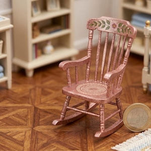 May include: Miniature pink rocking chair with floral and circular designs. The chair is part of a nursery set, with a changing table, crib, and shelves in the background. A coin is next to the chair.