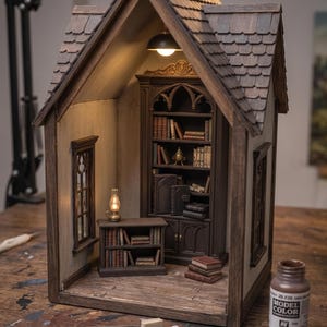 May include: Miniature wooden library diorama with a detailed interior. Features a bookshelf, small table, and books. The diorama has a shingled roof, a window, and a warm interior light. A bottle of Model Color paint is visible.