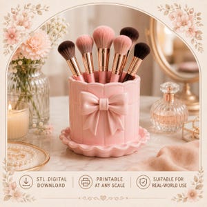 Pode incluir: Um suporte cilíndrico para pincéis de maquilhagem em rosa claro, com um laço moldado proeminente na parte frontal e uma base recortada. O recipiente contém vários pincéis com virolas em ouro rosa e cerdas macias e bicolores.
