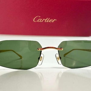 Peut inclure: Lunettes de soleil sans monture avec des verres verts et des branches en métal doré ornées d'une tête de panthère au niveau des charnières. Le logo Cartier est imprimé sur le verre supérieur gauche. Les lunettes sont devant une boîte rouge Cartier.