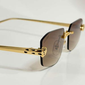 Peut inclure: Lunettes de soleil sans monture avec des verres dégradés marron et le logo Cartier imprimé sur le verre droit. Les branches en métal doré sont ornées d'un motif panthère noir tacheté au niveau des charnières, reliées à de fines branches dorées.