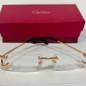Peut inclure: Lunettes sans monture avec des branches et un pont en métal doré, dotées de verres transparents et d'embouts de branches noirs. Les lunettes sont placées devant une boîte rectangulaire rouge de la marque Cartier avec une inscription dorée.