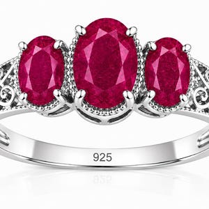 May include: A silver ring featuring three oval-cut deep red gemstones set in a row. The central stone is larger than the two flanking stones. The band displays intricate metal scrollwork patterns on both sides and is stamped with 925.