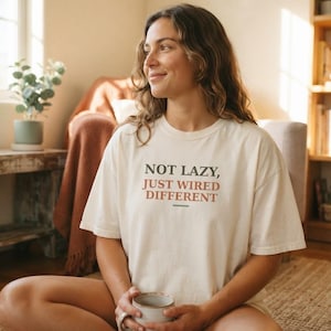 Puede incluir: Una camiseta de manga corta en color blanco roto con el texto "NOT LAZY, JUST WIRED DIFFERENT" impreso en letras de color verde oscuro y terracota en el pecho, acompañado de un pequeño gráfico de línea horizontal debajo.