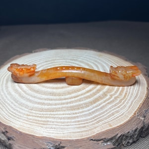 May include: A carved, orange-toned jade ornament. The piece features a curved design with a dragon head at each end. The ornament is supported by a small, rectangular base and rests on a wooden surface.