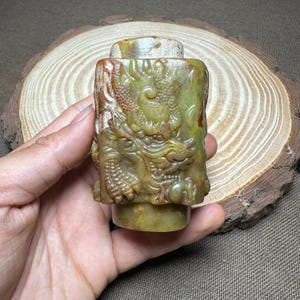 May include: A carved, rectangular jade object with a detailed dragon design. The jade is primarily green with brown and tan accents. The dragon is intricately carved, showcasing scales and facial features. The object has a cylindrical base and a flat top.