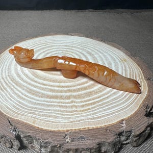 May include: A carved, orange-toned jade dragon figurine. The dragon is depicted in a curved pose, with detailed scales and a head facing left. The figurine is displayed on a wooden slice with visible growth rings.