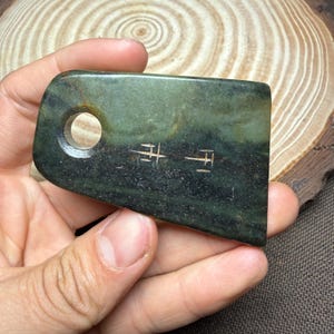 Op de afbeelding: Een donkergroene jade steen met een rechthoekige vorm en een afgeronde hoek. Het heeft een gat in de buurt van de bovenkant en geëtste tekens. De steen wordt in een hand gehouden, met een houten achtergrond.