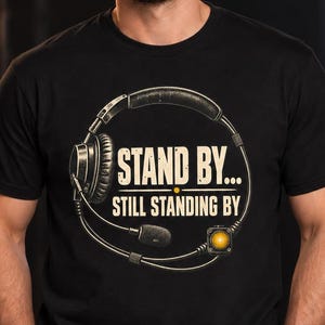 Puede incluir: Una camiseta negra de manga corta con un gráfico de auriculares circulares con micrófono y una luz indicadora amarilla. El texto "STAND BY... STILL STANDING BY" está impreso en el centro del gráfico de los auriculares en una fuente clara y negrita.