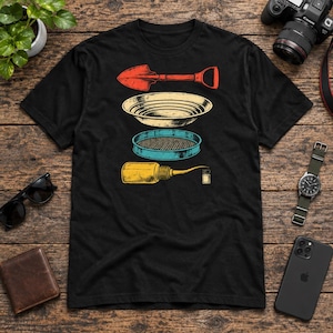 May include: A black t-shirt featuring a graphic print of gold prospecting equipment. The design includes a red shovel, a beige gold pan, a blue sifter, a yellow squeeze bottle, and a small glass vial arranged in a vertical column.