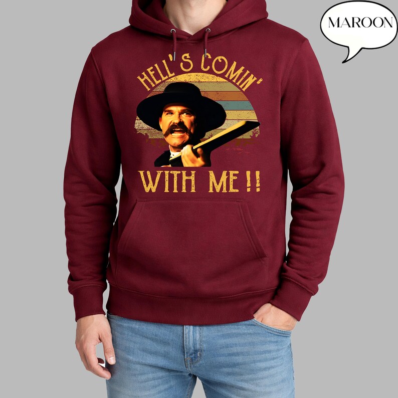 May include: A maroon hooded sweatshirt featuring a graphic print of a man with a mustache wearing a wide-brimmed hat. The text reads HELL'S COMIN' WITH ME!! above and below the figure, set against a circular background of horizontal stripes.