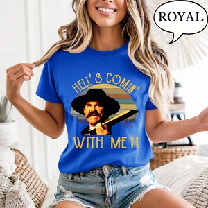 May include: A royal blue short-sleeve t-shirt featuring a graphic print of a man in a black hat with a mustache. The text reads HELL'S COMIN' WITH ME!! above and below the image, set against a retro-style sunset background in muted colors.