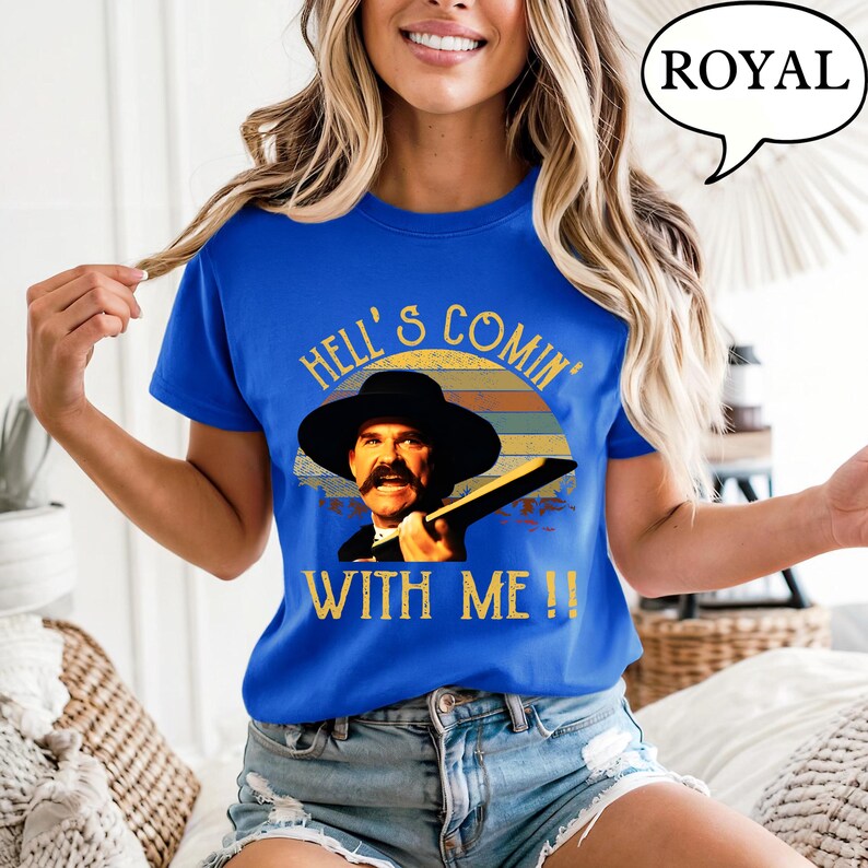 May include: A royal blue short-sleeve t-shirt featuring a graphic print of a man in a black hat with a mustache. The text reads HELL'S COMIN' WITH ME!! above and below the image, set against a retro-style sunset background in muted colors.