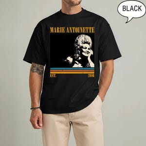 May include: Black t-shirt featuring a graphic of Marie Antoinette. The design includes the text "MARIE ANTOINETTE" in a gold font, a vintage-style image of Marie Antoinette, and the text "EST. 2006" in a retro font. The shirt is a classic crew neck.