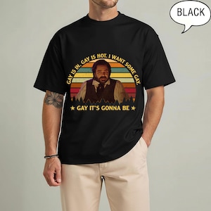 May include: A black t-shirt featuring a graphic print of a person in a vest. The design includes a sunset-colored background with the text: GAY IS IN. GAY IS HOT. I WANT SOME GAY. GAY IT'S GONNA BE. A speech bubble in the top right corner says BLACK.