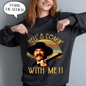 May include: A dark heather gray crewneck sweatshirt featuring a graphic print of a man with a mustache wearing a wide-brimmed hat. The text reads HELL'S COMIN' WITH ME!! above and below the image, set against a retro-style sunset background.
